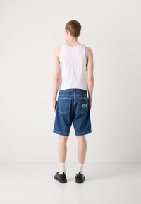 Vue arrière d'un homme portant un débardeur blanc, un short en jean bleu avec un logo à l'arrière et des chaussures noires avec des chaussettes blanches. Design simple, coupe décontractée.