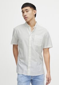 Camisa de manga corta con botones en tela gris clara, que presenta un único bolsillo en el pecho y un cuello, combinada con jeans de color azul claro.