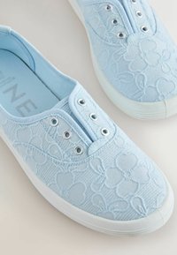 Baskets bleu clair à enfiler avec broderie florale et semelles en caoutchouc blanc, avec œillets métalliques sans lacets, posées sur une surface claire.