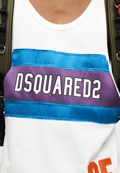 Débardeur blanc à rayures bleues et violettes avec le texte « DSQUARED2 », porté avec des bretelles de sac à dos vert foncé visibles sur les épaules.