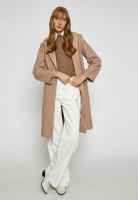 Hellbrauner langer Mantel mit einem strukturierten Strickpullover darunter, kombiniert mit weißen weiten Hosen und spitzen Schuhen.