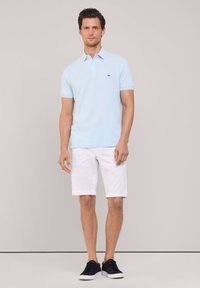 Polo shirt bleu clair avec un petit logo marine, associé à des shorts blancs. Modèle portant des baskets navy, se tenant contre un fond neutre.