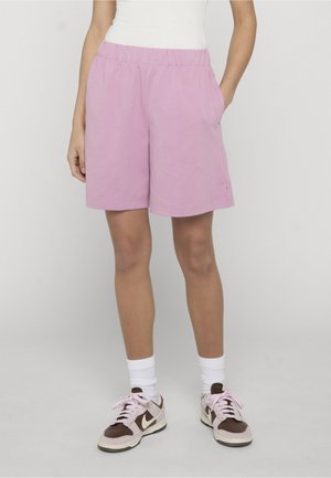 Person iført pastelfarvede lyserøde shorts, hvide sokker, brune og lyserøde sneakers og en hvid ærmeløs top, stående med den ene hånd i lommen.