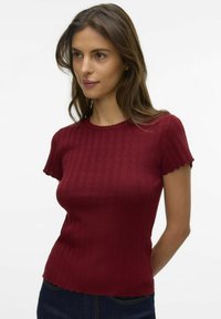 Haut en tricot ajusté bordeaux avec de courtes manches à volants, texture côtelée verticale et ourlet festonné, porté avec un jean en denim foncé.