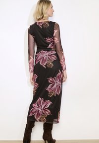 Žena v průhledných černých maxi šatech s dlouhým rukávem a velkými růžovými květinovými vzory, stojící zády k fotoaparátu, oblečená v tmavohnědých lodičkách na podpatku.