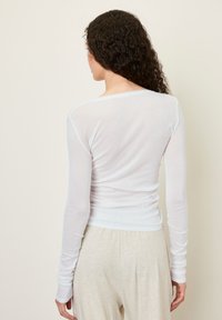 Top bianco a maniche lunghe con un design aderente, realizzato in un tessuto leggero e trasparente, caratterizzato da un collo rotondo e dettagli arricciati sul retro.
