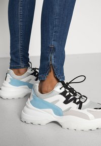 Sapatilhas chunky brancas e azul claro com sobreposições de malha e sintético, combinadas com jeans azul escuro com fechos éclair nos tornozelos.