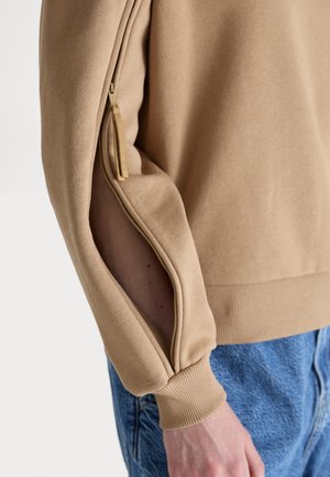 Beige hettegenser med glidelås på ermet og ribbestrikkede mansjetter, med en glatt tekstur og avslappet passform. Synlig sømdetalj forbedrer designet.