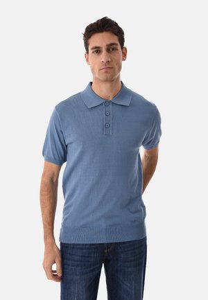 Uomo che indossa una polo azzurra a maniche corte con bottoni e jeans blu scuro, in piedi davanti a uno sfondo bianco uniforme.
