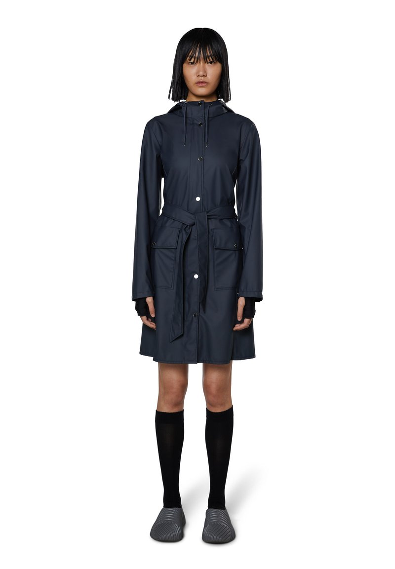 Tummansininen vedenpitävä trench-coat, jossa on huppu, kiristettävä vyötärö ja kaksi etutaskua. Varustettu napituspainikkeilla ja sileällä pinnalla. Yhdistä mustiin sukkiin ja teksturoituihin liukuviin kenkiin.