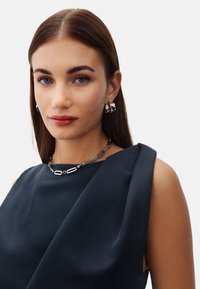 Svart satinklänning med draperad axel, silverlänkshalsband och matchande hoppringar, med släta texturer och en elegant design.