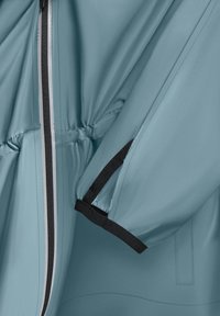 Veste imperméable en tissu doux de couleur sarcelle, équipée d'une fermeture éclair, d'une taille cintrée et d'accents en velcro noir sur les manches.