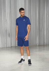 Blå trenings t-skjorte med rund hals og hvit logo, sammen med blå shorts med hvite sidestreker og svarte treningssko.