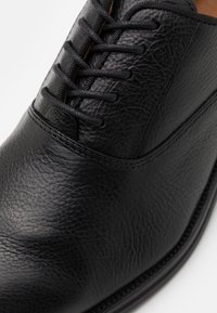 Chaussure en cuir noir à surface texturée, dotée de six œillets pour les lacets marron contrastants, avec des coutures détaillées le long du bord.