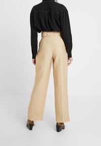 Osoba v béžových kalhotách s vysokým pasem a širokými nohavicemi, černém dlouhém tričku zasunutém dovnitř a hnědých vzorovaných botách na podpatku, stojící zády.