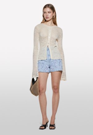 Donna che indossa un top crema in pizzo con bottoni, pantaloncini ricamati azzurri chiaro, sandali neri con tacco, che tiene una borsa intrecciata.