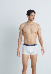 Weiße Boxershorts mit einem navyblauen Bund, auf dem der Markenname "GANT" steht. Der Stoff wirkt glatt und eng anliegend. Model steht vor einem weißen Hintergrund.