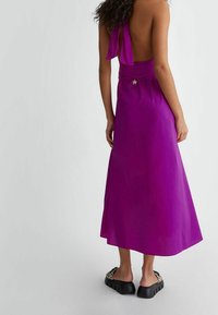 Fioletowa sukienka bez pleców z wiązaniem na szyi, zwiewny krój w kształcie litery A oraz gwiazda akcentująca w talii; noszona z czarnymi, masywnymi sandałami.