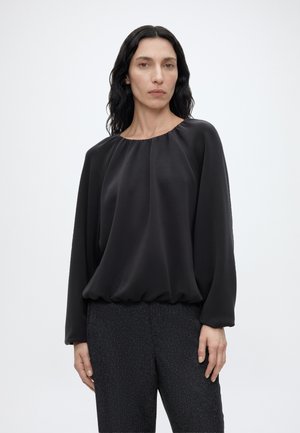 Femme aux longs cheveux noirs ondulés, portant un chemisier noir ample à manches longues et un pantalon foncé texturé, debout devant un fond clair uni.