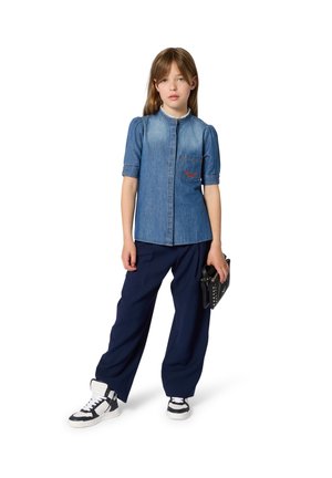 Fille portant un haut en denim à manches courtes, un pantalon large marine, des baskets noires et blanches, tenant une pochette noire cloutée, debout devant un fond blanc.