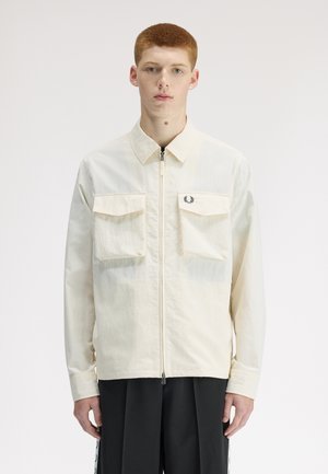 Jeune homme aux cheveux roux courts portant une veste beige à fermeture éclair avec poches avant et un pantalon noir, debout devant un fond blanc uni.