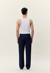 Moški stoji obrnjeno stran, oblečen v belo majico brez rokavov, zataknjeno v temno modre hlače in črne čevlje, na enostavni ozadju.