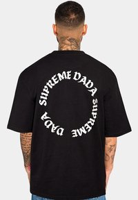 Schwarzes Baumwoll-T-Shirt mit einem geschwungenen wei ßen Textdesign, das auf dem Rücken "SUPREME DADA" liest. Lockere Passform mit kurzen Ärmeln.