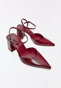 Sandales à talons blocs en cuir rouge, à bout pointu, avec des brides à la cheville et des boucles argentées sur fond blanc.