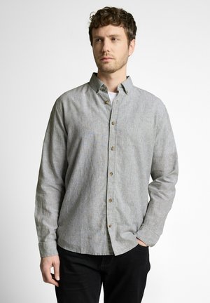 Man with light brown hair wearing a gray button-up shirt over a white undershirt, standing with one hand in pocket against white background.