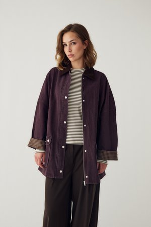 Kvinde iført en mørkelilla oversized jakke over en stribet langærmet trøje og brede sorte bukser, stående mod en ensfarvet lys baggrund.