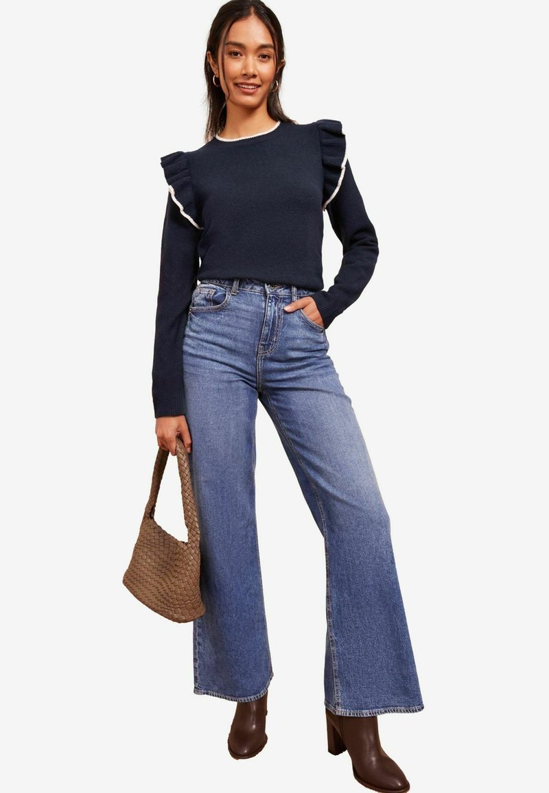 Pulover bleumarin cu accente volă pentru umeri, blugi albaștri cu talie înaltă și bocanci maro cu toc. Geanta din ratan maro are un design texturat.