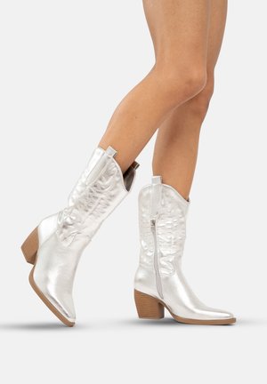Botas de vaquero metálicas plateadas con punta afilada, caña texturizada, cremallera lateral y tacones de madera apilados. Acabado suave y costuras decorativas.