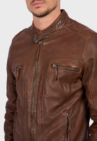 Giacca di pelle marrone con zip frontale, colletto alto e due tasche sul petto con zip. Presenta una superficie testurizzata e dettagli di cucitura.