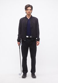 Άνδρας που στέκεται φορώντας μαύρο μπουφάν και παντελόνι, κρατώντας ένα γκολφ μπαστούνι, με μπλε σκούρο πουκάμισο και λευκή ζώνη, μπροστά σε απλό λευκό φόντο.