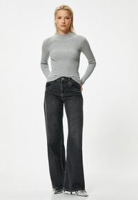 Grijze geribbelde lange mouwen top gecombineerd met een hoge taille, wijd uitlopende zwarte jeans met zakken en een knopen sluiting; model staat.