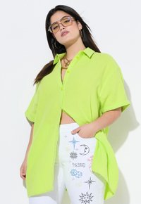 Camicia oversized verde neon con bottoni, realizzata in tessuto testurizzato, abbinata a pantaloni bianchi con disegni e motivi colorati stampati.