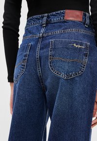 Džínsy z tmavomodrého denimu s vysokým pásom, ktoré majú dve zadné vrecká a hnedú koženú nášivku na opasku.