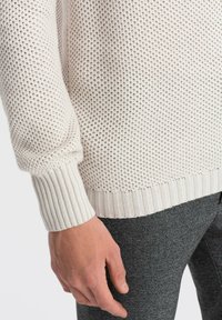 Pull en maille beige clair avec un tissage ouvert texturé, des poignets et un ourlet côtelés. Porté sur un pantalon à motifs gris foncé.