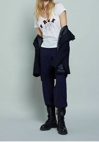 Bílý tričko s V výstřihem a červeným nápisem "FREE", spárované s tmavě modrými zúženými kalhotami a černými šněrovacími bojovými botami. Černý kabát přehodnocený přes ruku.