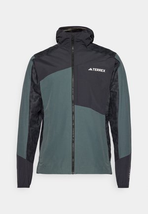 Lättvikts, vattentät jacka i mörkgrön och svart, med dragkedja fram, huva och ett mönstrat panel på ärmarna. Varumärkeslogotyp på bröstet.