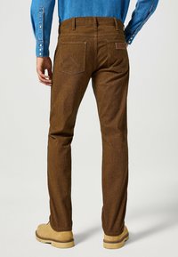 Pantalones marrones texturizados con bolsillos traseros y una etiqueta de marca de cuero. Botas de color claro visibles en la parte inferior. Camisa de mezclilla azul puesta.