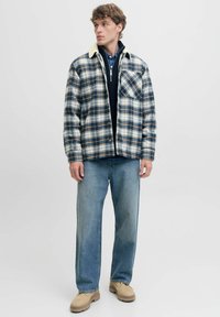 Veste à carreaux navy, crème et marron avec un col en polaire beige. Associée à un jean bleu clair et des bottes beiges. Une poche poitrine visible.