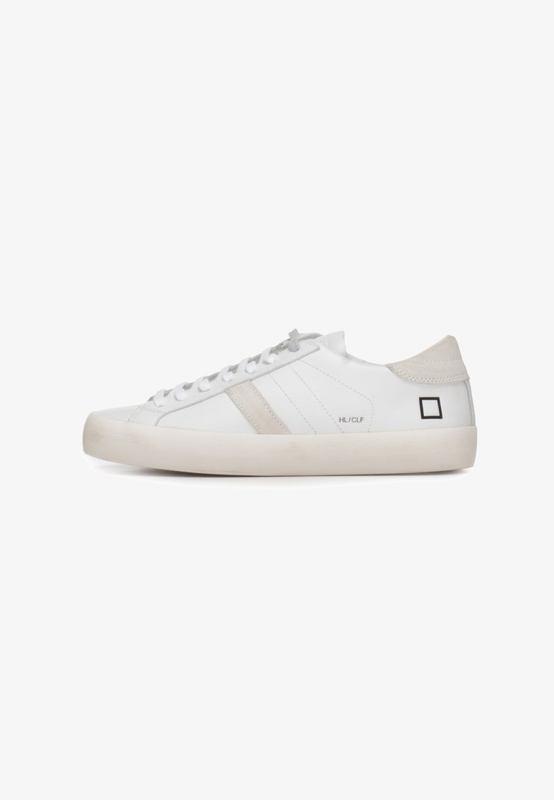 Witte lage sneaker met beige suède hielstuk, beige zijstreep, witte veters en zwart vierkant logo nabij de hiel op een witte zool.
