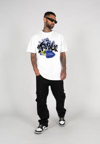 Hvid t-shirt med farverigt graffiti-inspireret design, der har en blå mund-illustration og sort skrift. Sorte cargo-bukser og sneakers.