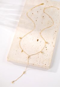 Naszyjnik łańcuchowy w odcieniu złotym, wielowarstwowy, z błyszczącymi, przezroczystymi kamieniami szlachetnymi, posiadający regulowaną długość i delikatne ogniwa, eksponowany na teksturowej powierzchni.