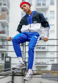 Jachetă de tip track în culorile negru, alb și albastru, cu manșete elastice; pantaloni albaștri asortați; adidași gri; fundal urban cu clădiri moderne.