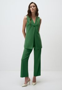 Gilet verde su misura e pantaloni a gamba larga realizzati in tessuto morbido. Il gilet presenta un bottone frontale e un design a strati; abbinato a scarpe con tacco alto color crema.