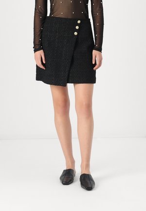 Mini-jupe en tweed noir avec un design à enroulement sur le devant, ornée de trois boutons dorés sur le côté. Associée à des mules plates noires.