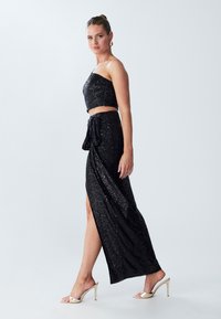 Černý flitrovaný dvoudílný outfit s bezramínkovým crop topem a maxi sukní s vysokým rozparkem, doplněný zlatými vysokými sandály.