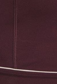 Tumši bordo audums ar vertikālām šuvēm un horizontālu baltu apdari tuvu apakšējai malai.